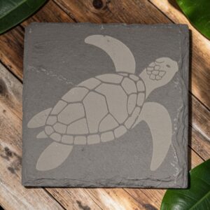 Square slate coaster with a laser-engraved Hawaiian honu sea turtle on charcoal-gray stone, displayed on a wooden table.