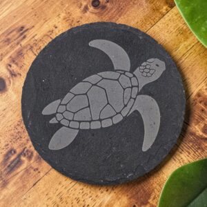 Round slate coaster with a laser-engraved Hawaiian honu sea turtle on dark charcoal stone, displayed on a wooden table.