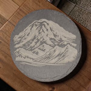 Round mountain slate coaster featuring detailed laser-engraved rugged peaks and rocky ridges on charcoal-gray stone.