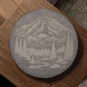 Round slate coaster with laser-engraved mountain peaks, evergreen forest, and winding river on natural charcoal-gray stone.