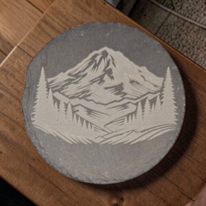 Round slate coaster with a laser-engraved mountain peak framed by evergreen trees on charcoal-gray stone.
