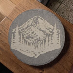 Round slate coaster featuring laser-engraved mountain peaks framed by evergreen trees on natural charcoal-gray stone.