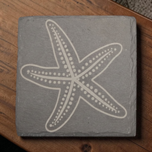 Square 4-inch charcoal slate coaster with a laser-engraved five-arm starfish and decorative dotted pattern on a wooden table.
